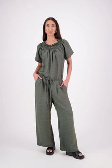 The N-E-S Muses Jumpsuit features a shirred off-the-shoulder top with cap sleeves, an elasticated back waist, and a self-drawcord front for a flattering, adjustable fit. The lower half mirrors the relaxed fit of our Lounge Jumpsuit, with angled seams, centre front detailing, and practical front pockets