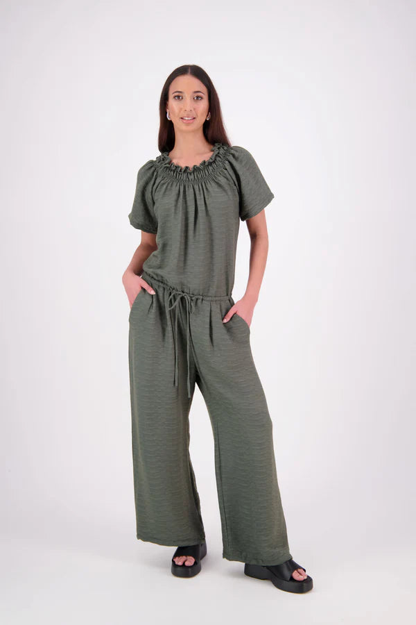 The N-E-S Muses Jumpsuit features a shirred off-the-shoulder top with cap sleeves, an elasticated back waist, and a self-drawcord front for a flattering, adjustable fit. The lower half mirrors the relaxed fit of our Lounge Jumpsuit, with angled seams, centre front detailing, and practical front pockets