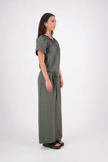 The N-E-S Muses Jumpsuit features a shirred off-the-shoulder top with cap sleeves, an elasticated back waist, and a self-drawcord front for a flattering, adjustable fit. The lower half mirrors the relaxed fit of our Lounge Jumpsuit, with angled seams, centre front detailing, and practical front pockets